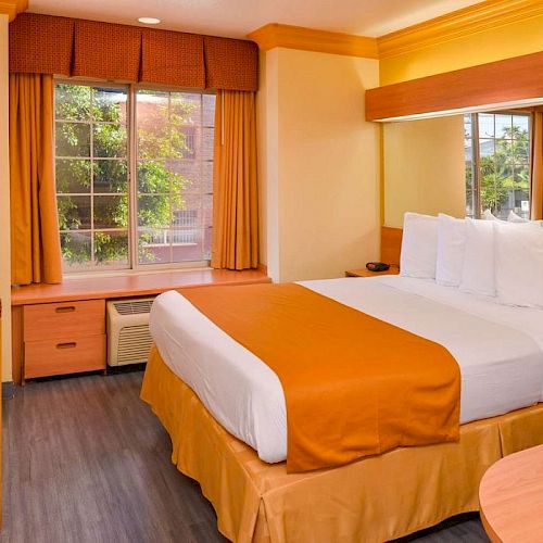 A cozy hotel room features a neatly made bed, a window with brown curtains, a desk with a chair, and a mirror above the headboard.