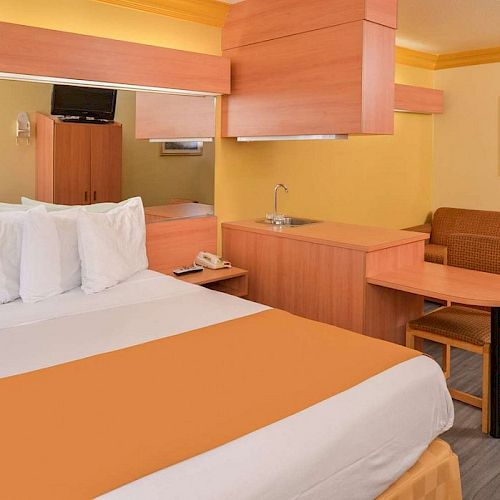 A cozy hotel room features a bed, desk, chair, small kitchenette, TV, sofa, and framed artwork on yellow and orange walls, and wooden flooring.