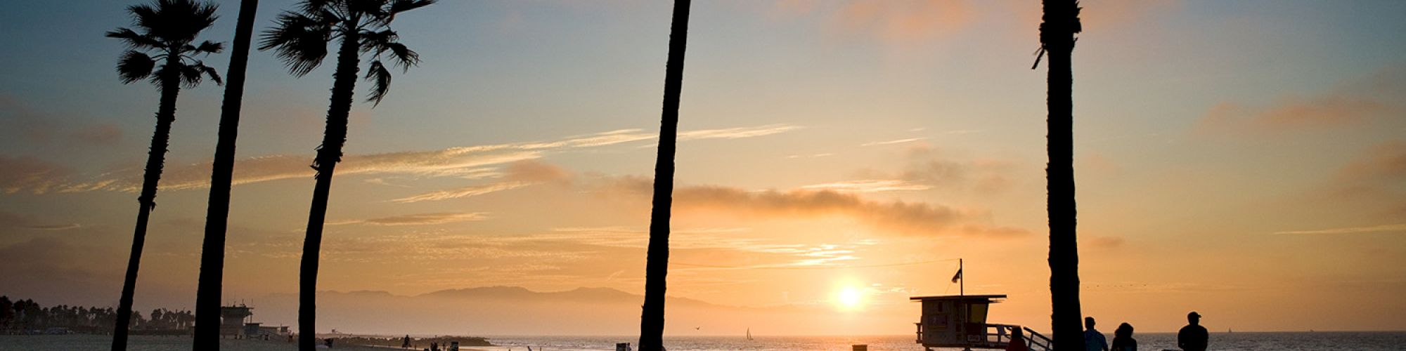 Sunset at a beach with tall palm trees, a lifeguard tower in the distance, and people enjoying the peaceful scene near the shoreline.