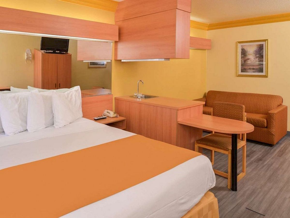 A cozy hotel room with a double bed, small couch, desk, TV, microwave, and wall art, all in warm yellow and wood tones.